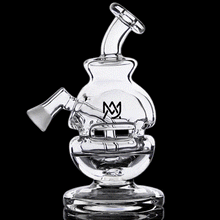 The MJ Arsenal Royale Vape Edition is a clear glass water pipe with a unique, artistic design, featuring a curved neck, angled mouthpiece, and black stylized M logo on its main chamber against a black background.