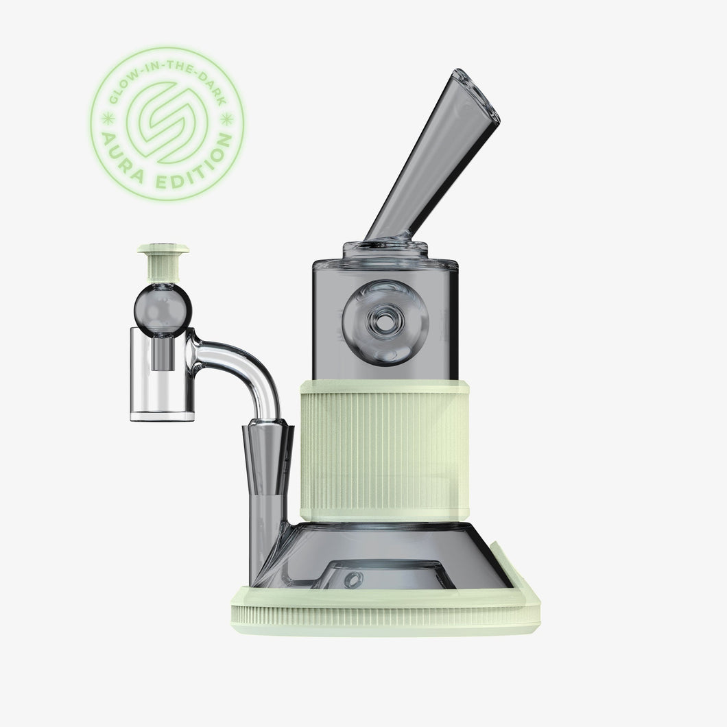 The SOFTGLASS AURA TANDEM RIG is a modern electronic dab rig with a compact design, glow-in-the-dark white accents, an angled mouthpiece, and attached glass bubbler—plus optional silicone sleeves—shown on a plain white background.