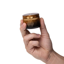 A hand holds the Human Grade Grinder 7F (2.5"), a small, round gold and black aluminum herb grinder with a geometric pattern, against a plain white background.