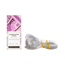 A purple and white box labeled Grav Sandblasted Spoon sits beside a deep bowl pipe wrapped in bubble wrap with yellow rubber bands. The packaging showcases illustrations of smoking accessories.