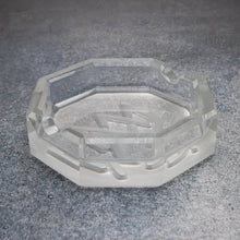 The MJ Arsenal Pegasus Ashtray, a clear octagonal luxury piece with geometric faceted edges, sits on a textured gray surface. Its reflective base catches the light, creating elegant patterns and highlights.