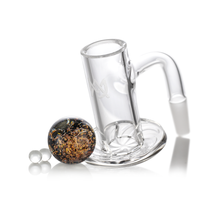 The MJ Arsenal Full Weld Quartz Terp Blender is a clear glass accessory with a cylindrical chamber, angled mouthpiece, flat base, two small glass beads, and a round cap with brown and gold flecks—ideal for use with MJ Arsenal Mini Rigs.