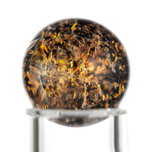 A close-up of the MJ Arsenal Full Weld Quartz Terp Blender shows a glass sphere on a clear stand, filled with swirling golden and brown flecks. Its abstract design stands out against the plain white background.