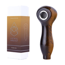 A brown Gemstone Spoon Pipe with a polished finish and geometric white designs on a gradient background rests beside its Astral Project Tiger Eye box. This unique pipe features a metal bowl and striking gemstone craftsmanship.