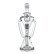 The MJ Arsenal Time Turner Mini Dab Rig is a clear, hourglass-shaped rig with intricate, symmetrical glasswork and a central cone chamber, standing upright on a reflective surface.