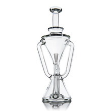 The MJ Arsenal Time Turner Mini Dab Rig is a clear borosilicate glass rig shaped like an hourglass with a top stopper, two symmetrical handles, and is shown on a reflective surface with a white background.
