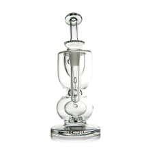 The MJ Arsenal Titan Mini Dab Rig is a clear borosilicate glass piece featuring a cylindrical chamber, thick base, and central vertical handle-like detail, shown against a white background.