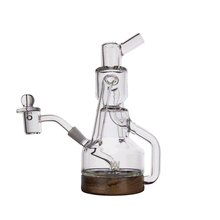 The MJ Arsenal Alpine Series - Apex Mini Rig is a clear glass water pipe with a handle, cylindrical chamber, angled mouthpiece, detachable bowl, and compact double uptake recycler design, shown standing on a round wooden base against a white background.