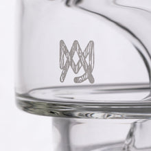 Close-up of the MJ Arsenal Alpine Series - Apex Mini Rig, with a white geometric crown logo etched on its clear surface. Its transparent glass and double uptake recycler design stand out against a plain white background.
