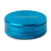 The Human Grade Grinder 1C is a blue, round, 2.5" aluminum grinder with textured sides, "HUMAN GRADE" printed in white on top, and a sturdy 2-piece design.