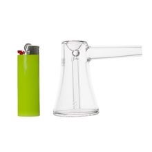 A green lighter stands next to the MJ Arsenal Vulkan Bubbler, a portable borosilicate glass piece with a wide base and straight mouthpiece. Both are upright on a white background.