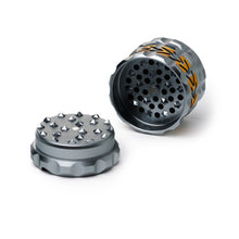 The Human Grade Grinder 8AT is a 4-piece aluminum grinder with sharp teeth, a built-in pollen screen, and an inner grinding compartment. It has a gray finish accented by orange flame-like designs on the side.
