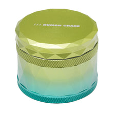 The Human Grade Grinder 7F (2.5") is a faceted, cylindrical aluminum herb grinder featuring a green-to-blue gradient design and "HUMAN GRADE" printed on the lid.