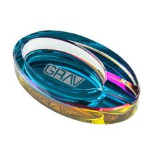 The Grav Ellipse Ashtray is a vibrant, oval-shaped iridescent tray with "GRAV" etched in the center, featuring blue, yellow, and pink hues plus a groove for resting a cigarette or cigar.