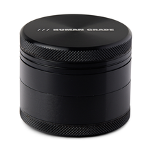 The Human Grade Grinder 1A (2" 4-Piece) is a black, cylindrical aluminum grinder with textured edges and "HUMAN GRADE" in white on the lid, featuring a built-in pollen screen for easy collection.