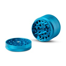 The Human Grade Grinder 1A (2" 4-Piece) in blue aluminum is shown with the lid off, revealing sharp grinding teeth, filtering holes, a pollen screen, and a textured grip. All parts are displayed on a clean white background.