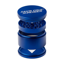 A blue Santa Cruz Shredder Large 4-Piece Grinder is shown open and upright, displaying its grinding teeth, mesh screen, and collection chambers. The brand name appears in white on the top lid.