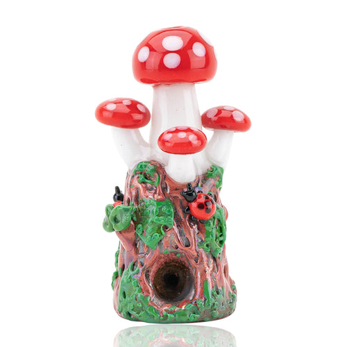 The Empire Glass Works Mushrooms Dry Pipe is a hand-crafted ornament shaped like red and white mushrooms growing from a tree stump, adorned with green leaves, vines, two red ladybugs, and features a circular hole at the base.