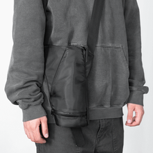 A person in a dark gray hoodie and black pants carries the Puffco Journey Bag—with a vertical zipper, ideal for Peak Pro accessories—over their shoulder, set against a plain light background.
