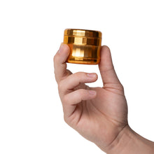 A hand holds the Human Grade Grinder 6M (2.5"), an ergonomic, gold-colored, textured aluminum herb grinder with a cylindrical design, shown against a white background.