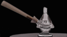 A wooden mallet strikes the MJ Arsenal King Bubbler, a pyramid-shaped borosilicate glass piece with a spout and engraved logo, shattering it on its circular base against a black background.