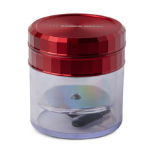 The Human Grade Storage Grinder 1A (2.5") features a clear cylindrical container with a textured metallic red lid labeled HUMAN GRADE. An aluminum grinder with a pollen screen is visible inside through the transparent sides. White background.
