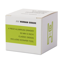 The Human Grade Grinder 1A (2" 4-Piece) comes in a white box labeled HUMAN GRADE, featuring a classic aluminum grinder with pollen screen, scraper tool, and a green line illustration on the side. Size: 50 mm (2 inch).