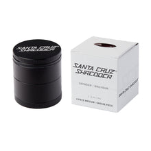 A black Santa Cruz Shredder Medium 4-Piece Grinder sits beside its white box, which displays the brand name and product details boldly in black text.