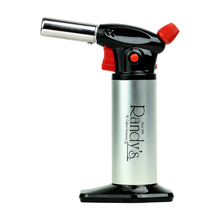 Randy's Torch is a silver and black butane torch with an ergonomic design, red ignition and adjustment knobs, and a black base. The brand “Randy’s” is printed on its side.