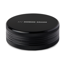 The Human Grade Grinder 1C (2.5" 2-Piece) is a black, round aluminum grinder with textured sides and "HUMAN GRADE" in white on top, offering a sturdy and stylish way to prepare your herbs.