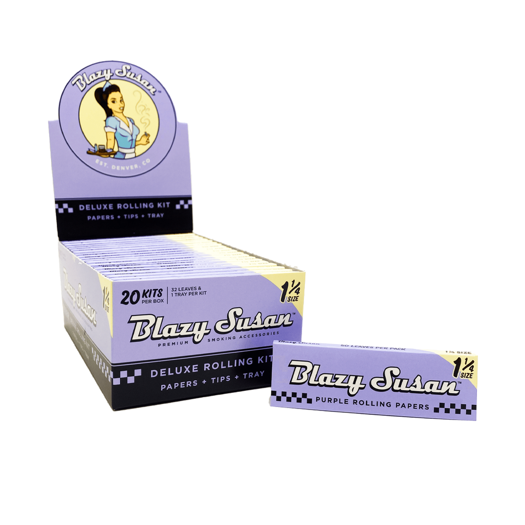 A box display of Blazy Susan Purple Rolling Papers 1 1/4 size, featuring vibrant purple papers, tips, and a tray. One individual deluxe rolling kit is shown in front of the packaging.