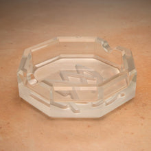 The MJ Arsenal Pegasus Ashtray is a clear, octagonal luxury ashtray with four cigarette grooves, shown on a light brown, textured surface.