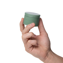 A hand holds the Human Grade Grinder 4C (2.5"), a small, round, green aluminum herb grinder with a gray lid, isolated on a white background.