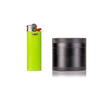 A bright green Bic lighter stands upright next to a black MJ Arsenal 4 Piece Grinder with a textured grip, both on a white background.